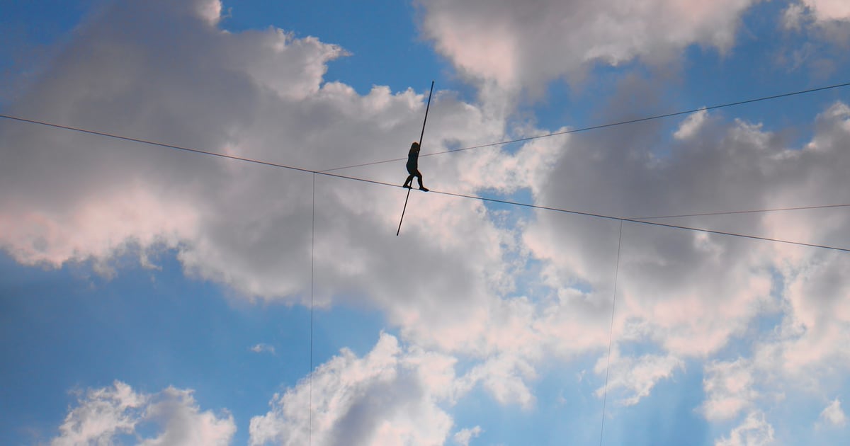 A small figure walking a slackline strung high across a sky of scattered clouds