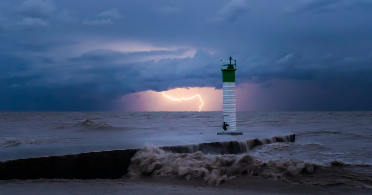 A white lighthouse on a breakwater as waves hit the pier under a dark sky lit by lightning