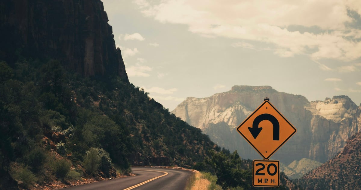 A yellow U-turn warning sign with a 20 mph plate beside a road that curves through a desert canyon