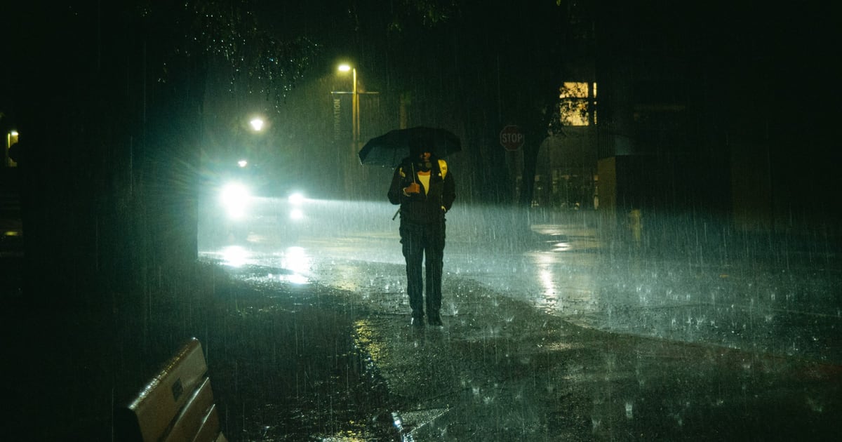 A person with an umbrella walking along a wet street at night in heavy rain, car headlights behind