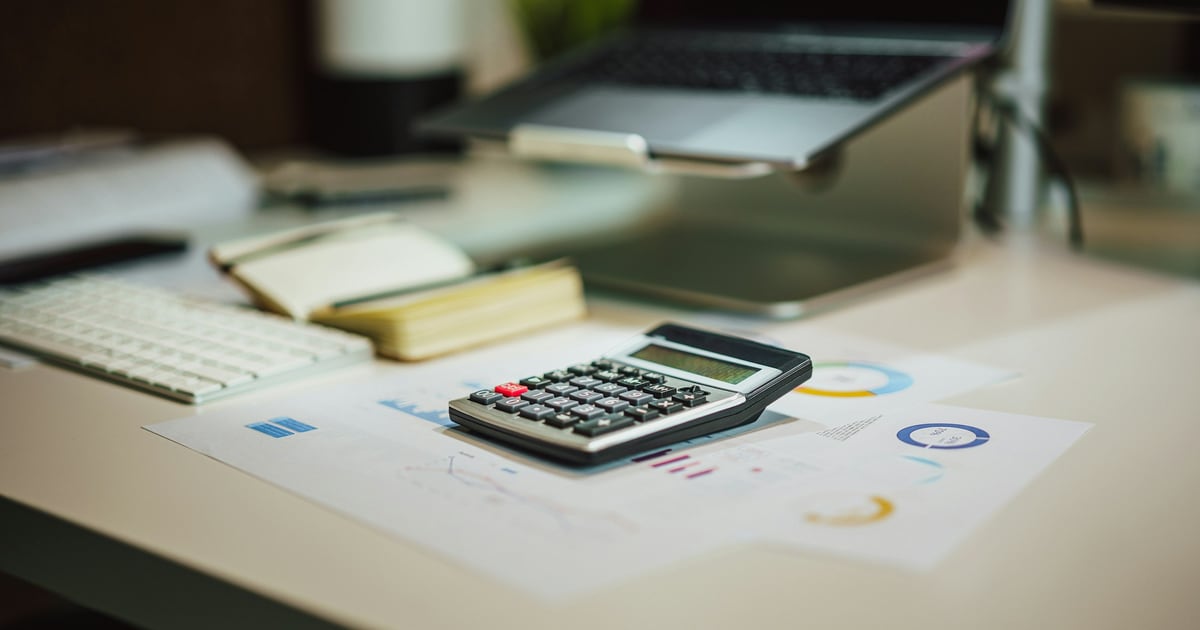 A desk with a calculator lying on printed charts, beside a keyboard, a notebook and a laptop on a stand