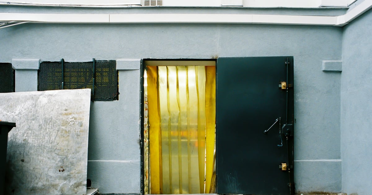 An open steel vault door in a pale blue wall, with gold-colored strips hanging across the doorway