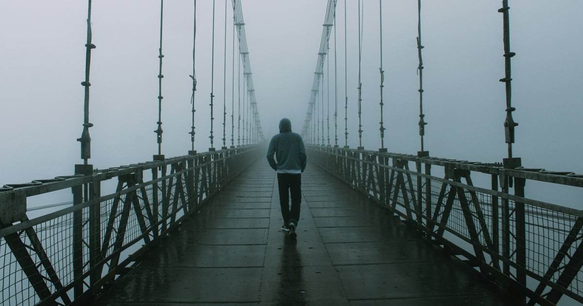 A person in a hooded jacket walking alone across a suspension footbridge that disappears into fog