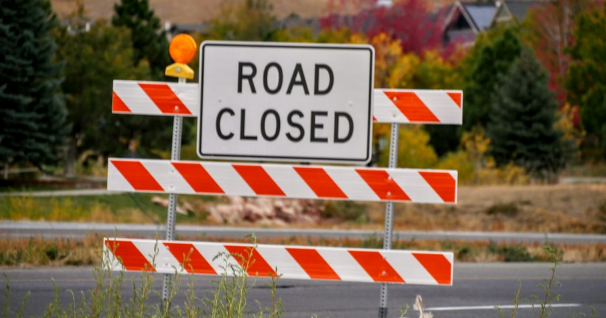 A Road Closed sign on striped orange and white barriers across a road, with trees behind